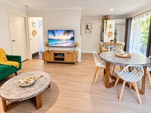 A modern living room with a flat-screen TV, a green sofa, a dining area with a round table and chairs, and a kitchen area with a window.