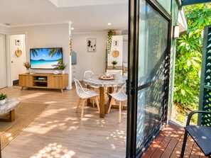 A modern living room with a TV, dining table, and chairs, a wooden cabinet, and a glass door leading to a balcony with greenery.