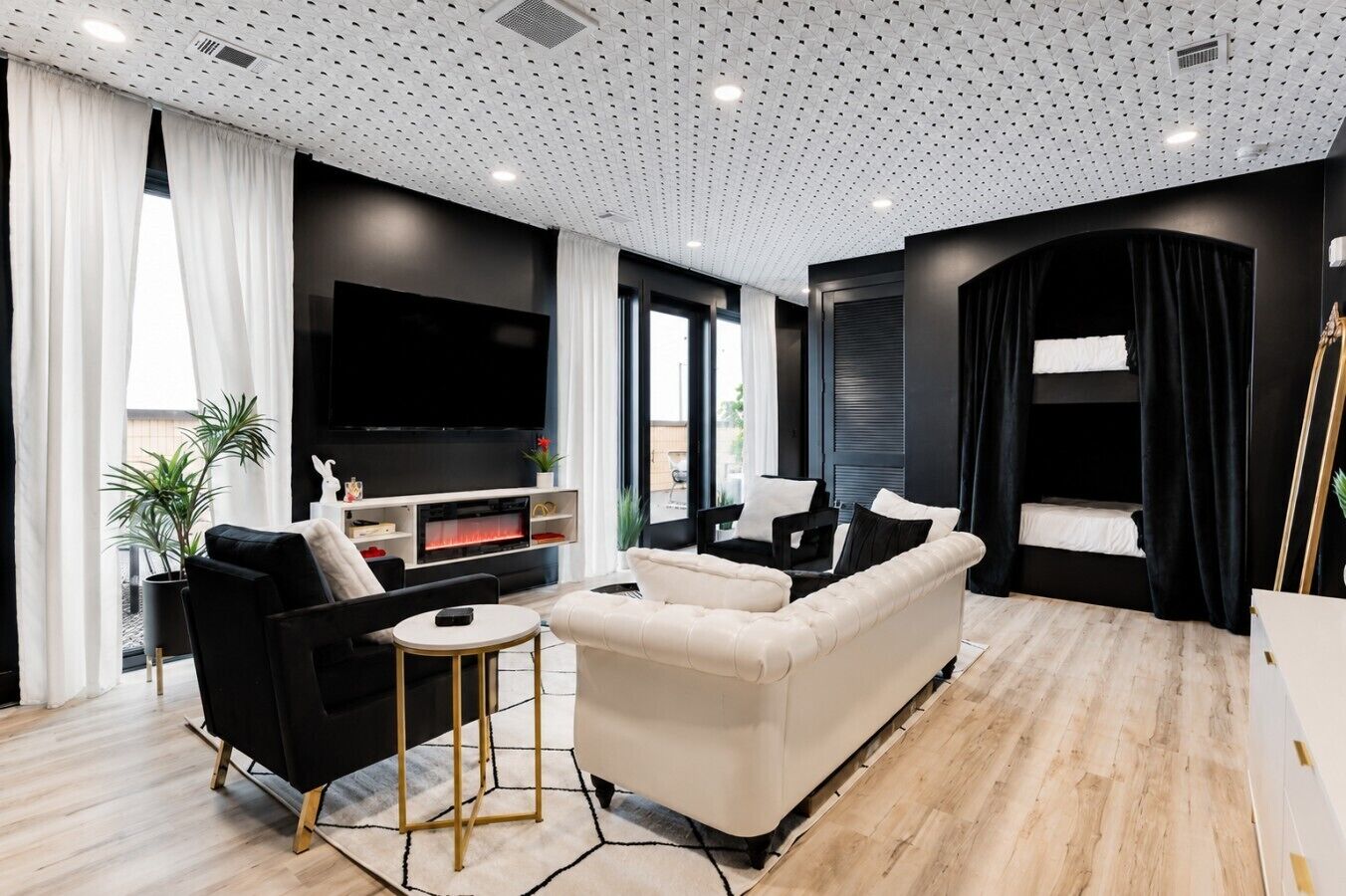 A modern living room with a flat-screen TV, black and white sofas, a small round table, and a large window with curtains.