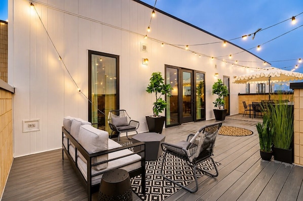 Un patio exterior con un sofá, sillas, una mesa, luces colgantes y plantas en macetas.