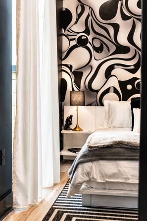 Une chambre à coucher avec un lit, une table de chevet, une lampe et un mur orné d’un motif abstrait en noir et blanc.