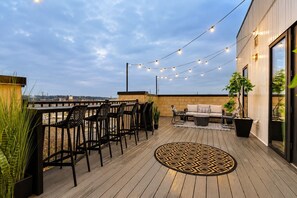 A rooftop terrace with a wooden deck, outdoor seating, string lights, and potted plants.