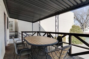 Een overdekt terras buiten met een ronde tafel en stoelen, een balustrade en uitzicht op gebouwen en bomen.