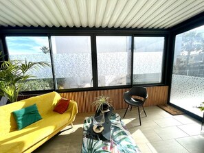 Um quarto claro com um sofá amarelo, uma cadeira preta e uma mesa com uma planta. Janelas amplas proporcionam vista para o exterior.