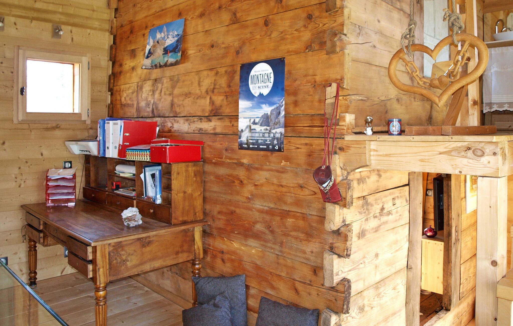 A rustic wooden cabin interior with a wooden desk, shelves with books and binders, a poster of Montagne en Scène, and a wooden heart-shaped decoration.