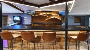 Angolo bar con un bancone in legno, quattro sgabelli marroni, un grande schermo a parete e una decorazione murale a tema montagna.