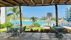 Un área de estar al aire libre techada, con mesas y sillas, con vista a una piscina y la playa.