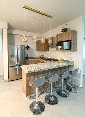 A modern kitchen with a central island, stainless steel appliances, hanging pendant lights, and bar stools.