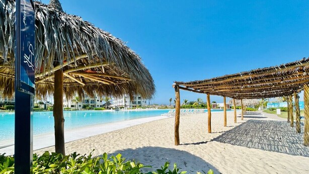 Una playa con piscina, sombrillas de paja y un área de descanso cubierta.