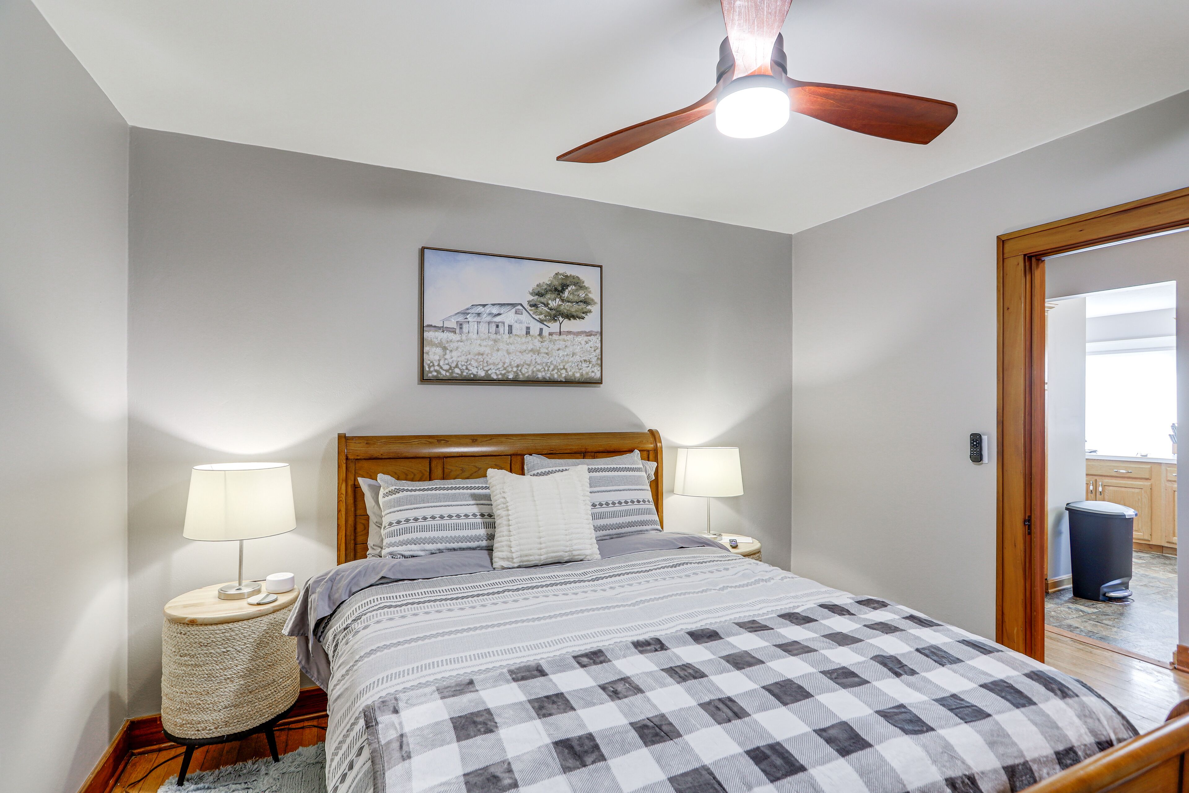 A bedroom with a bed, two bedside lamps, a ceiling fan, a framed picture on the wall, and a door leading to a bathroom.