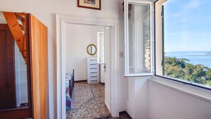 Un couloir donnant sur la mer, un escalier en bois, un lit recouvert d’une courtepointe colorée et une commode blanche.