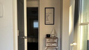 A hallway with wooden flooring, a door on the left, a wall-mounted sign, and a small white dresser with a decorative item on top.