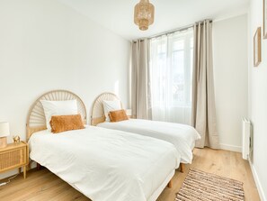 Two single beds with wooden headboards and white bedding, set against a white backdrop with a large window and woven rug.