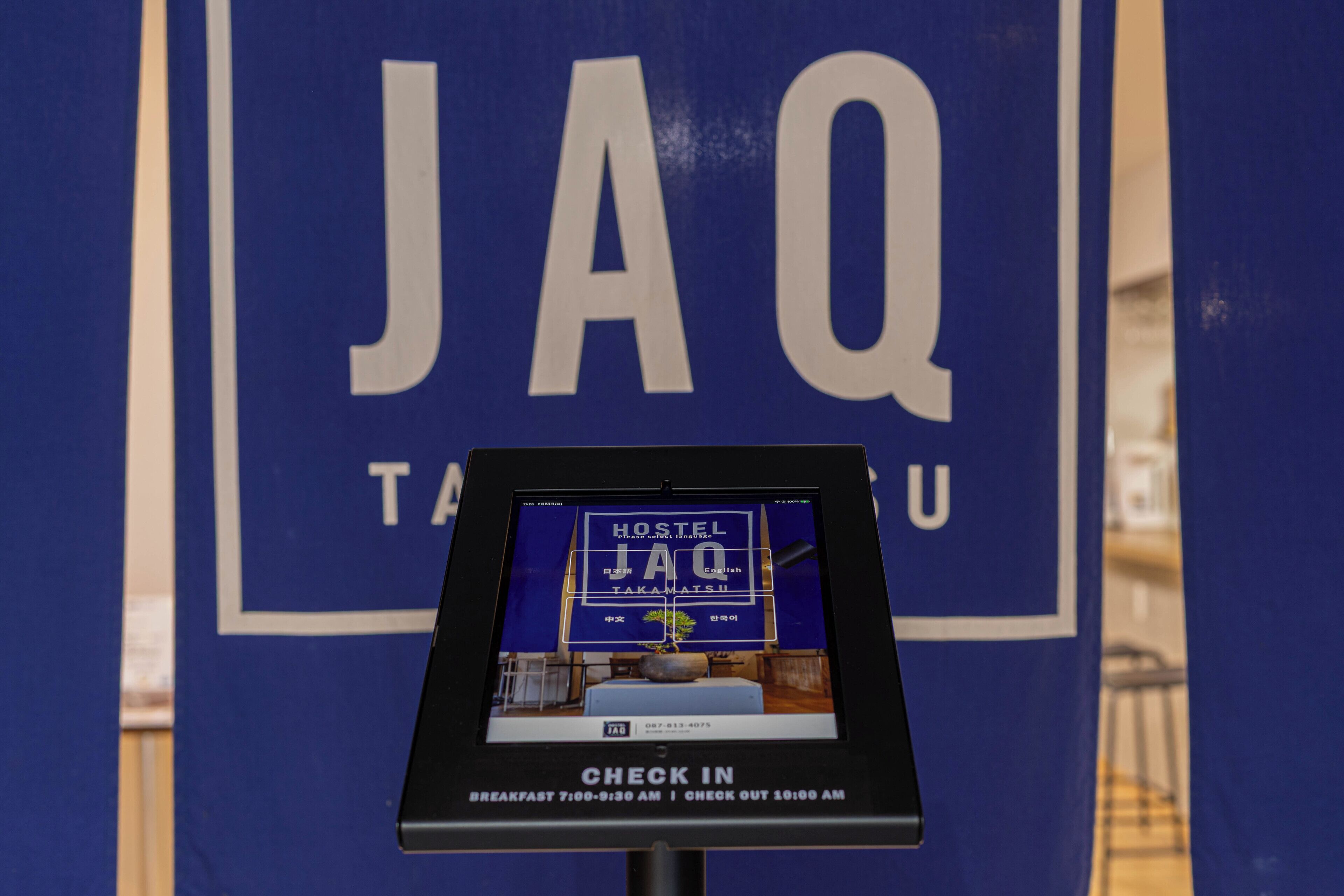 A digital kiosk with a Hostel JaQ advertisement, indicating check-in times and contact number.