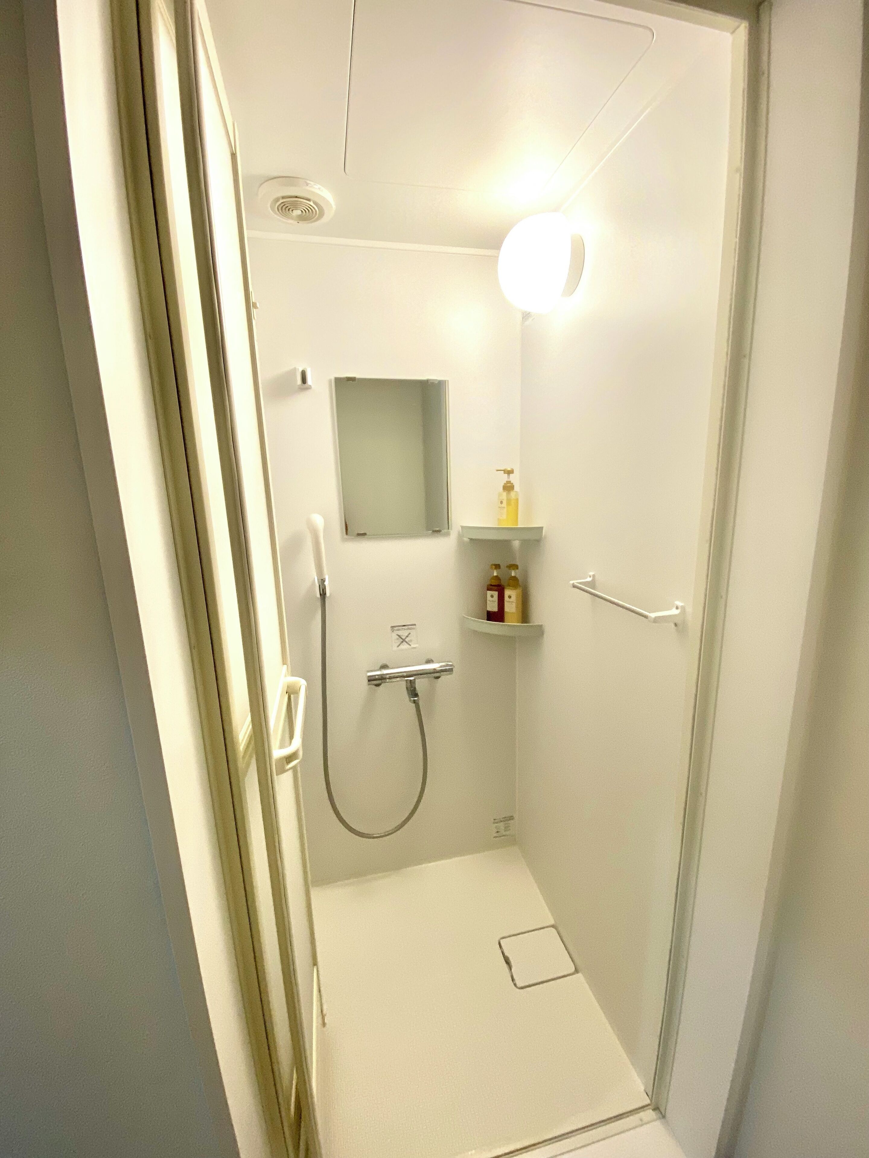 Un pequeño box de ducha blanco con cabezal de ducha de mano, un espejo montado en la pared y un estante con artículos de aseo.