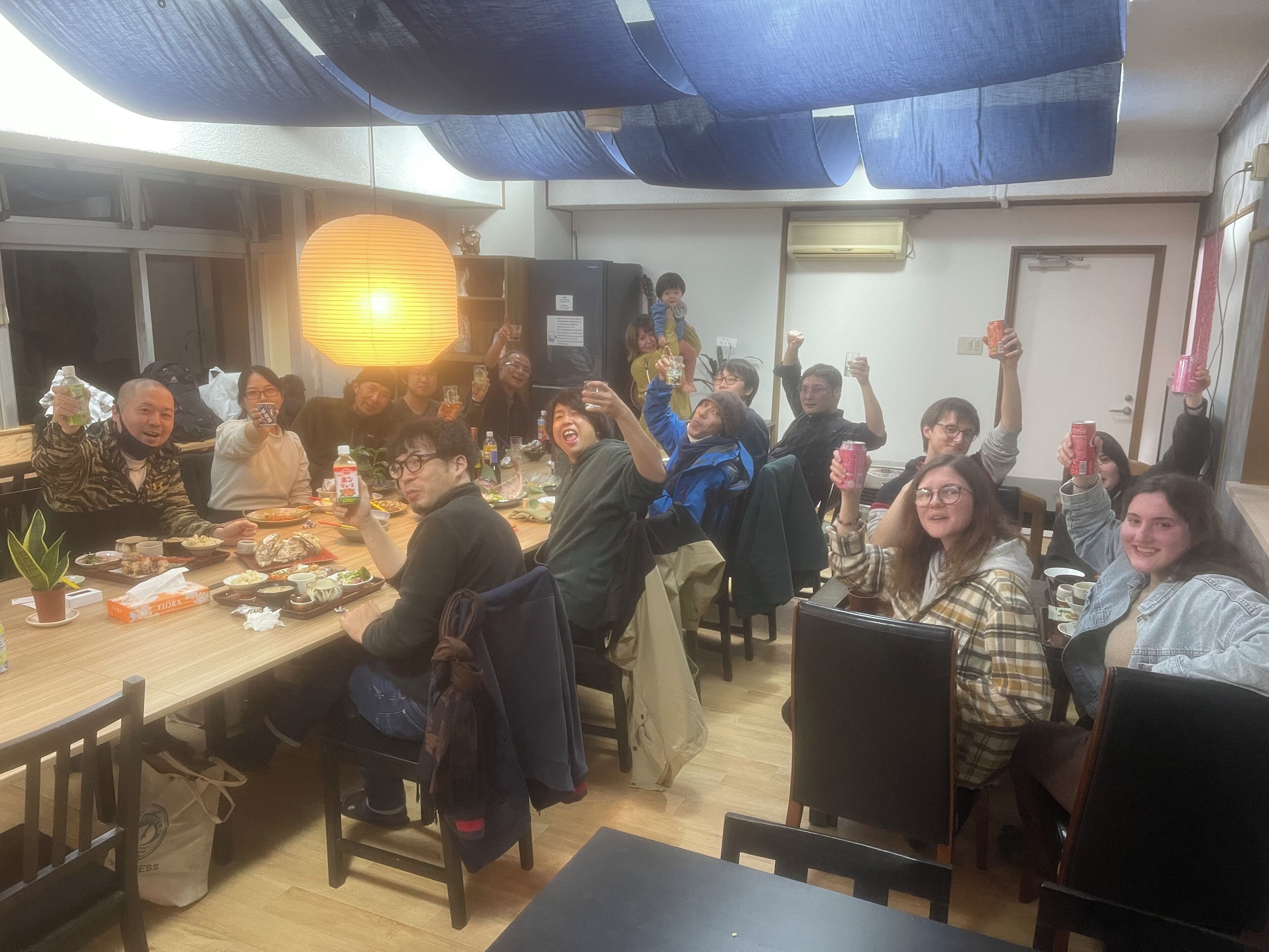 A group of people gathered around a table in a room with a wooden floor, a hanging light fixture, and a door in the background.