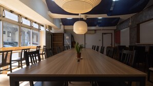A conference room with a long table, chairs, a potted plant, and a hanging light fixture. - Hostel JAQ takamatsu (Takamatsu)