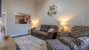 A living room with a brown leather sofa, patterned armchair, and a coffee table. There is a decorative wall piece above the sofa and a lamp on each side.