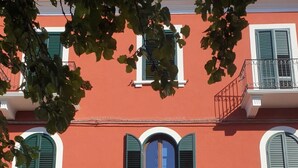 A building with a red facade, white trim, and green shutters. There are balconies with wrought iron railings and a large central door.