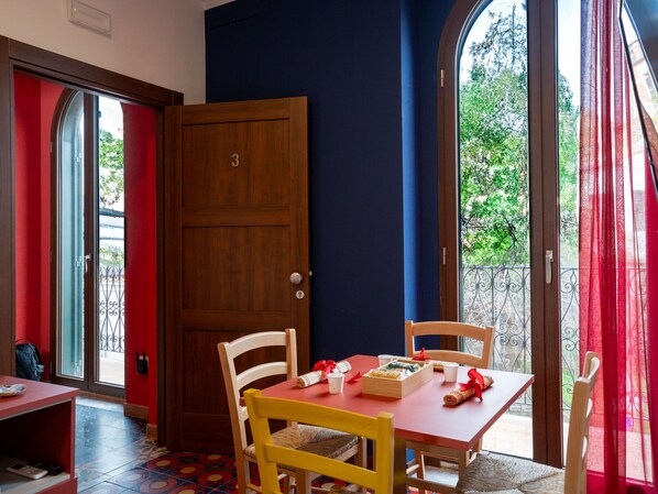 Une pièce avec une porte en bois, une table rouge avec des chaises, et une vue sur la verdure à l’extérieur.