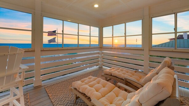A sunlit room with large windows, white furniture, and a view of the ocean at sunset. - PELICAN PLACE: Gulf Front 75ft Private Beach! Pool + Hot Tub! Elevator, Outdoor Kitchen, Green Egg (Inlet Beach)