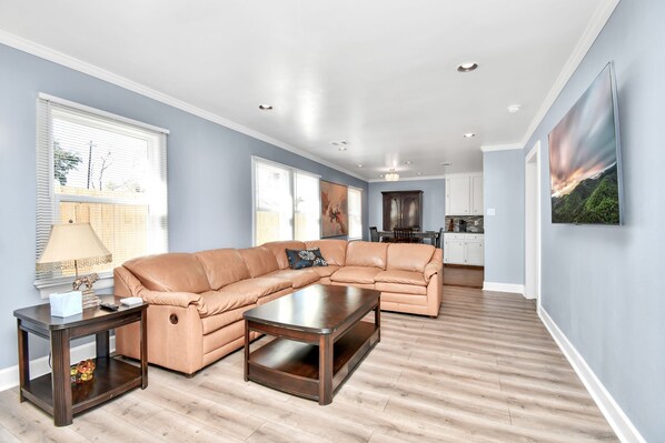A living room with a sectional sofa, a coffee table, and a side table. There is a kitchen area in the background with cabinets and a dining table. - Family Friendly - Clean & Spacious, Near Downtown NRG/Med CTR (Houston)