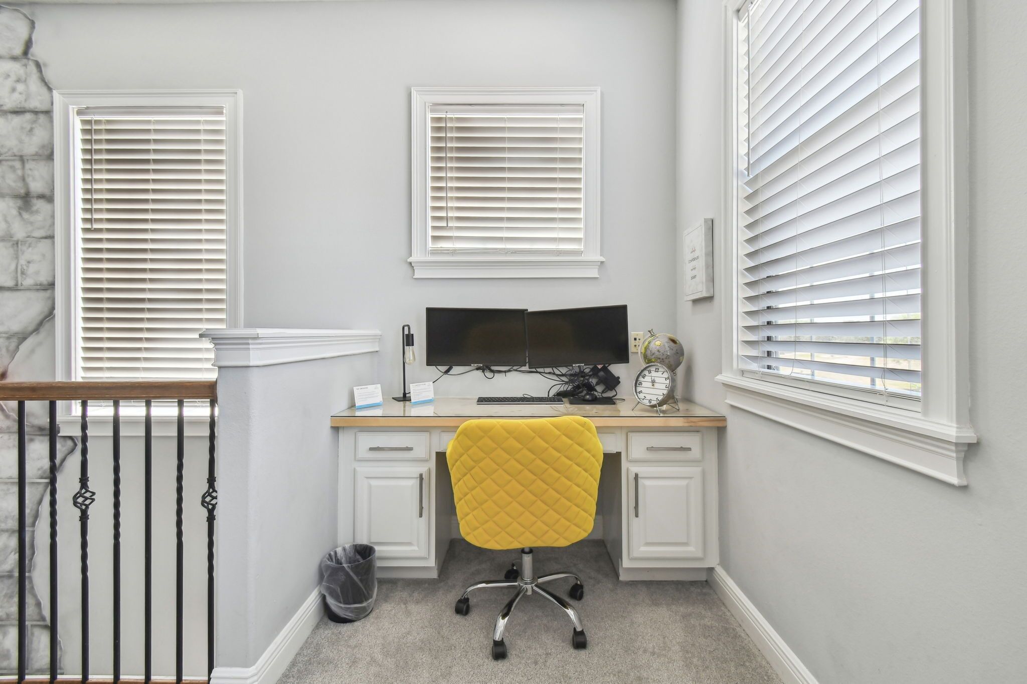 A study with a desk, computer, and a yellow chair. There are two windows with blinds, a stone wall, and a staircase.