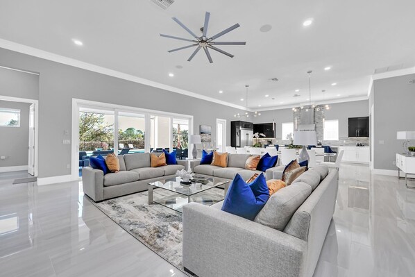 A spacious living room with a modern ceiling fan, a large glass coffee table, and grey sofas with blue and orange cushions. - Revive at Reunion | 10BR Luxe Villa w/ Private Pool, Theater, Arcade, and Themed Rooms Near Disney (Kissimmee)
