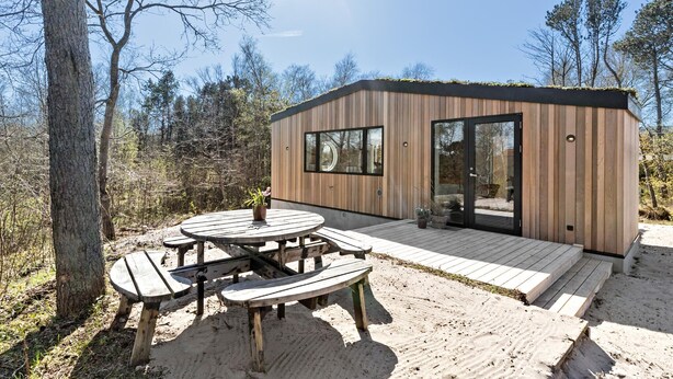 En modern stuga i trä med en veranda och picknickbord, omgiven av träd.