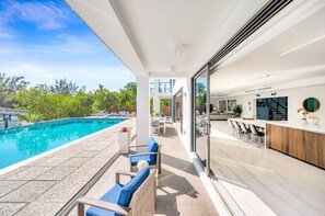 Un área de piscina moderna al aire libre con sillones de descanso, un comedor con bar y vistas a la piscina y al follaje.