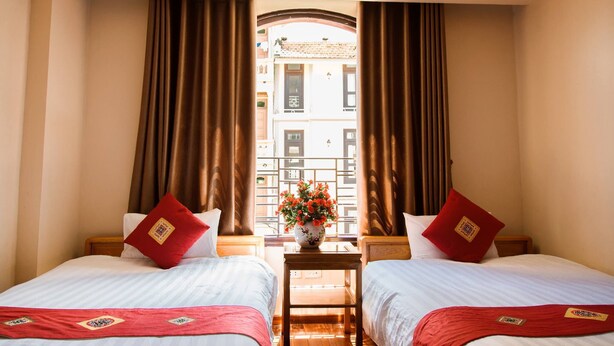 A hotel room with two beds, a small table with a vase of flowers, and a view of a building through a window with curtains. - Thu Hang Sapa Hotel - by Bay Luxury (Sa Pa)
