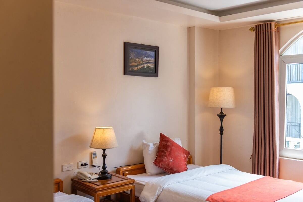 A hotel room with a bed, two bedside lamps, a nightstand with a telephone, a painting on the wall, and a window with curtains.