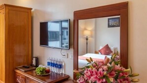 A hotel room with a wooden wardrobe, a desk with a laptop, water bottles, a fruit bowl, and a mirror reflecting a bed with a lamp. - Thu Hang Sapa Hotel - by Bay Luxury (Sa Pa)