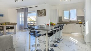 A modern kitchen with a dining area, featuring a large table with bar stools, a refrigerator, and a window with curtains.