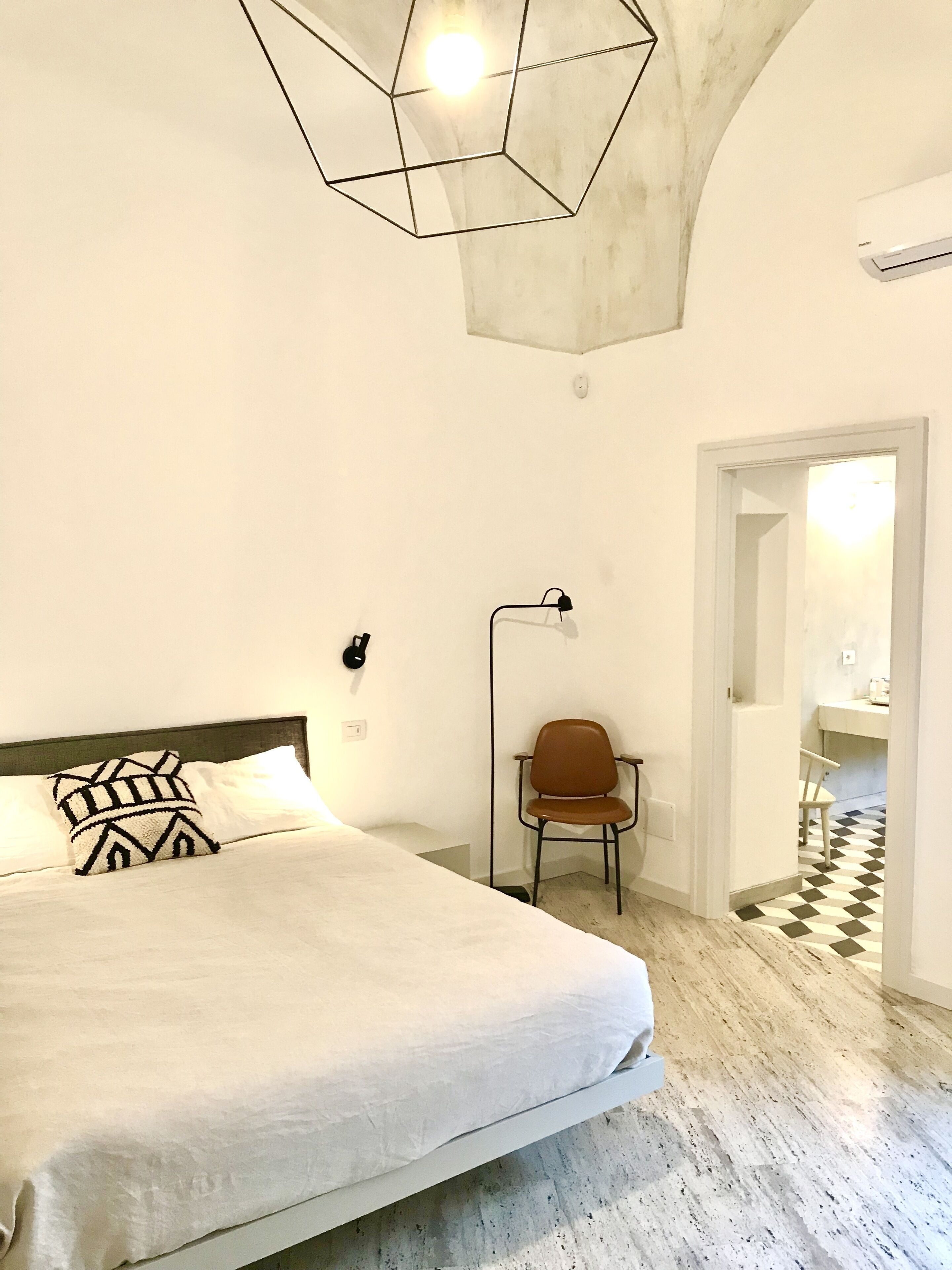 A bedroom with a large bed, a chair, a desk, and a bathroom visible through an open door.