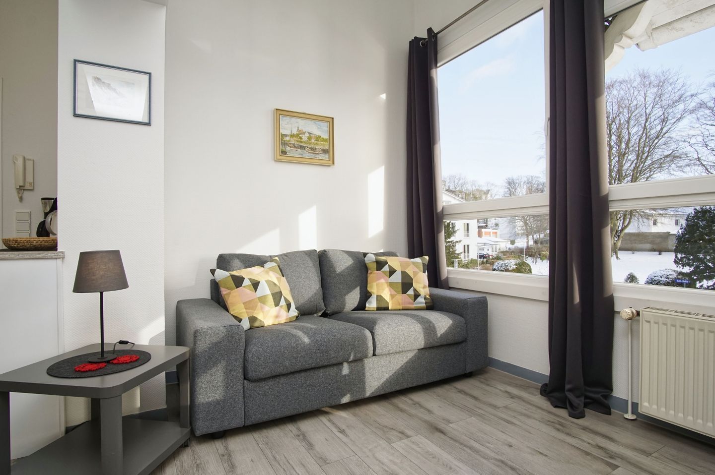 A modern living room with a grey sofa, two patterned cushions, a side table with a lamp, and a window with curtains.