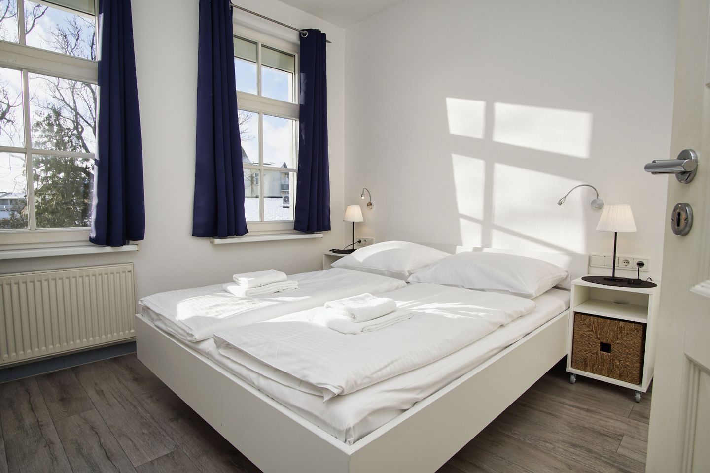 A neatly made bed in a room with a window, a bedside table with a lamp, and a radiator.