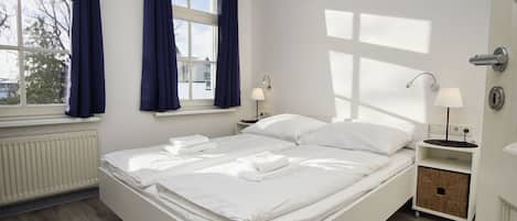 Ein ordentlich bezogenes Bett in einem Zimmer mit Fenster, Nachttisch mit Lampe und Heizkörper.