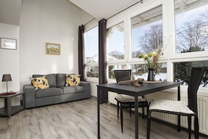 A modern living room with a grey sofa, a dining table with chairs, and a large window with a view of a snowy landscape.