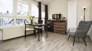 A modern living room with a dining table, two chairs, a grey armchair, a TV on a wooden cabinet, and a view of buildings through large windows. - im Zentrum Gelegene 2-raum-wohnung - Haus Rugier Fewo 03 - Haus Rugier (Göhren)
