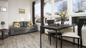 A modern living room with a grey sofa, a dining table with chairs, and a large window with a view of a snowy landscape. - im Zentrum Gelegene 2-raum-wohnung - Haus Rugier Fewo 03 - Haus Rugier (Göhren)