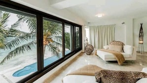 Un salon moderne doté d’une grande fenêtre donnant sur une piscine et des palmiers, d’un canapé blanc et d’une lampe décorative.