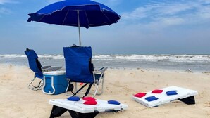 A beach setup with a blue umbrella, two beach chairs, a cooler, and shuffleboard tables. - Gulf Views, Walk to Sandfest, Shared Pools, Boardwalk to the Beach (Port Aransas)