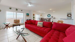 A spacious living area with a red sofa, a glass coffee table, and a striped armchair. There is a dining area with a table and chairs in the background. - Gulf Views, Walk to Sandfest, Shared Pools, Boardwalk to the Beach (Port Aransas)