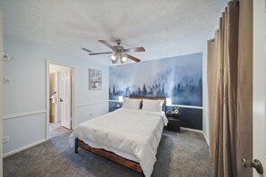Un dormitorio con cama, ventilador de techo, mesita de noche, un mural en la pared y una puerta.