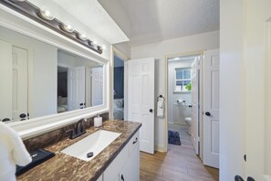Un baño con un espejo grande, un lavabo y una puerta que da a otra habitación.