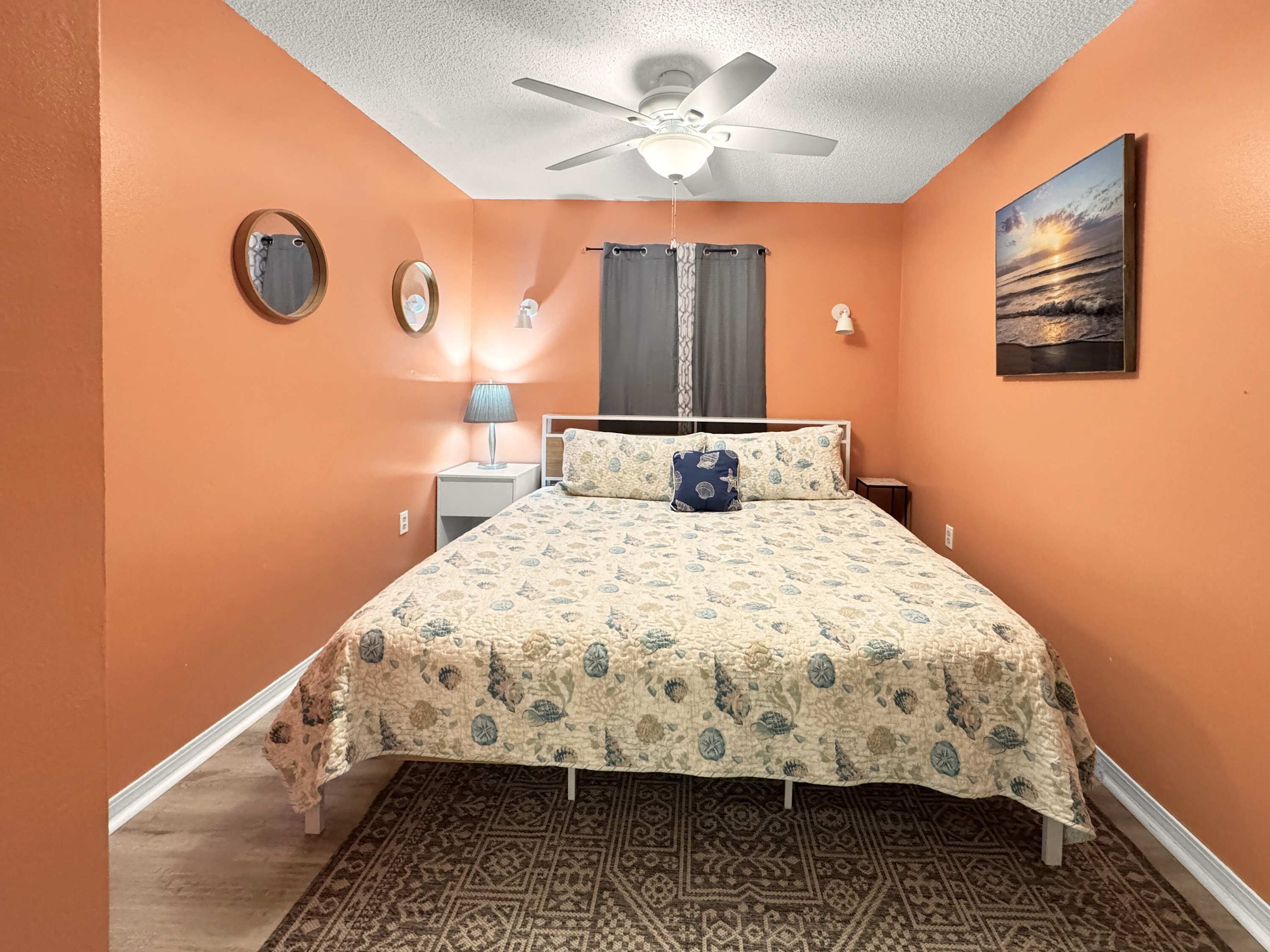 A bedroom with an orange wall, a bed with a patterned bedspread, a ceiling fan, a nightstand with a lamp, a mirror, a window with curtains, and a painting of a sunset.