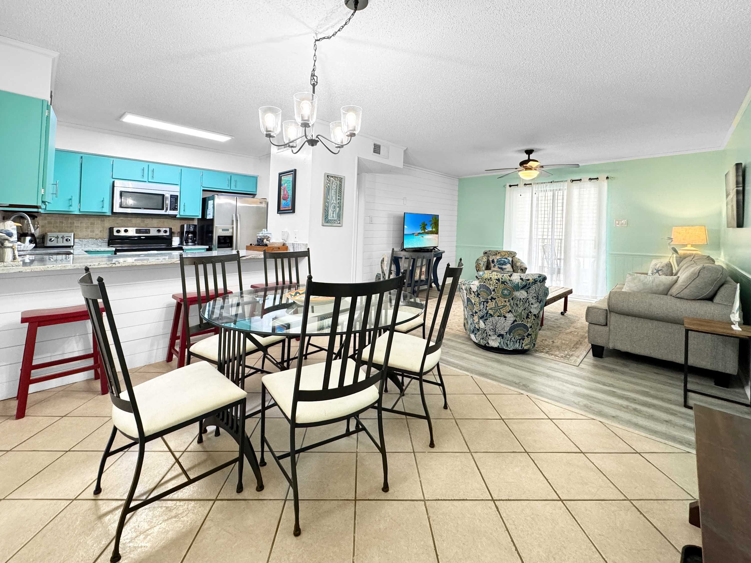 A modern kitchen with teal cabinets, a dining area with a glass table and black chairs, and a living room with a sofa and a TV.