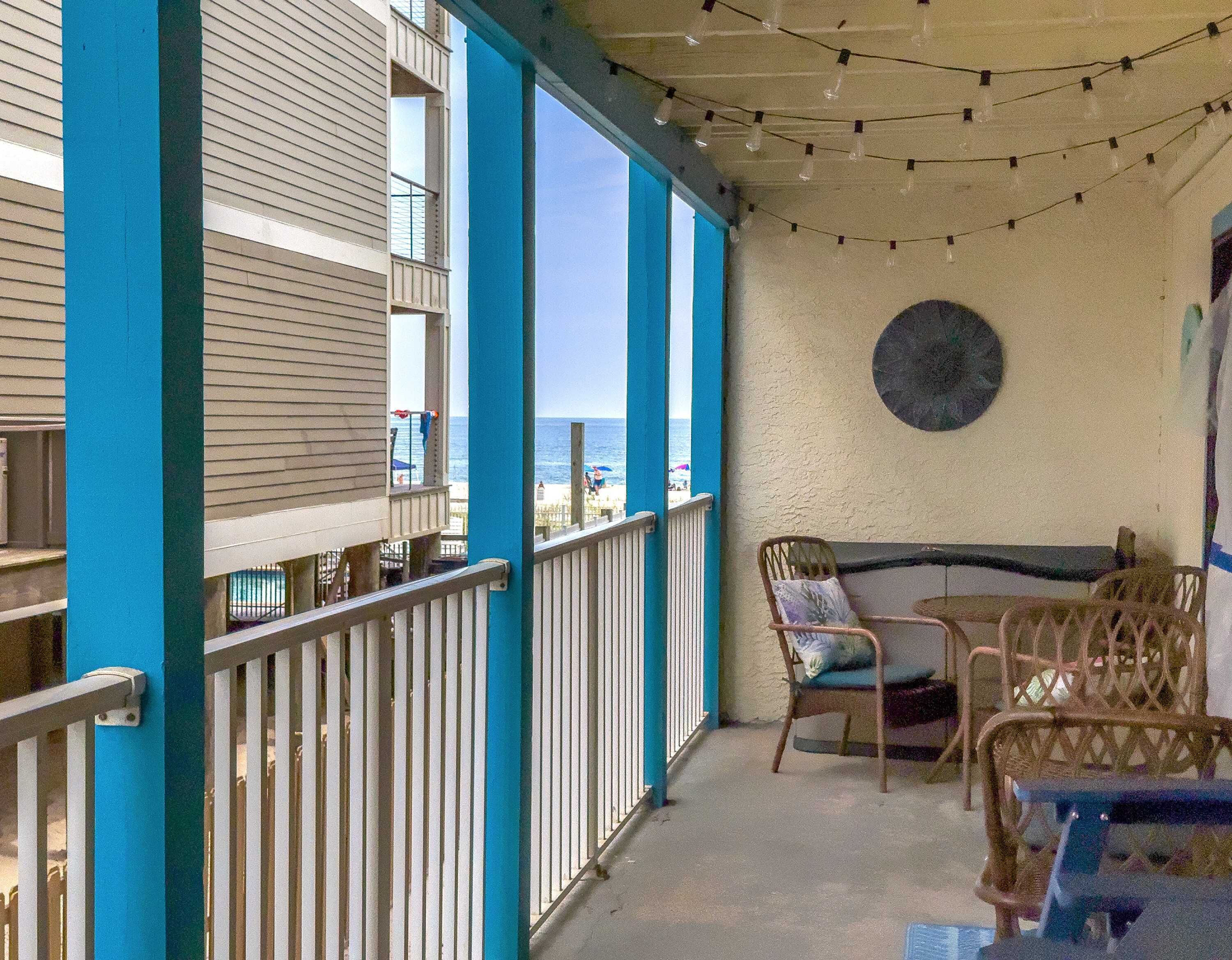 A covered outdoor area with a view of the beach, featuring a table and chairs, a round table, and a wall-mounted clock.