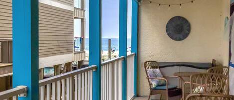 A covered outdoor area with a view of the beach, featuring a table and chairs, a round table, and a wall-mounted clock.